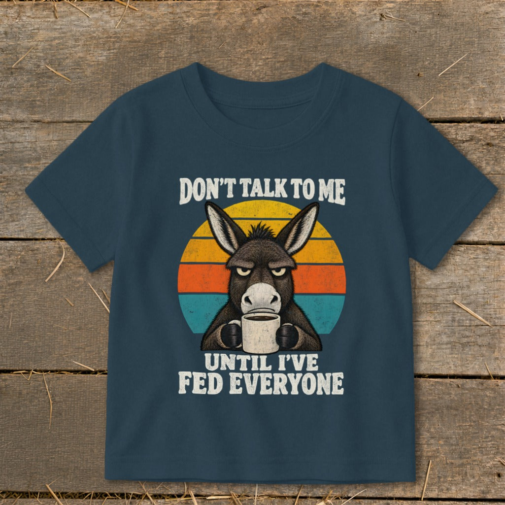 Dark blue t-shirt featuring a grumpy donkey holding a coffee mug, with text saying "DONT TALK TO ME UNTIL IVE FED EVERYONE" and a retro sunset graphic.
