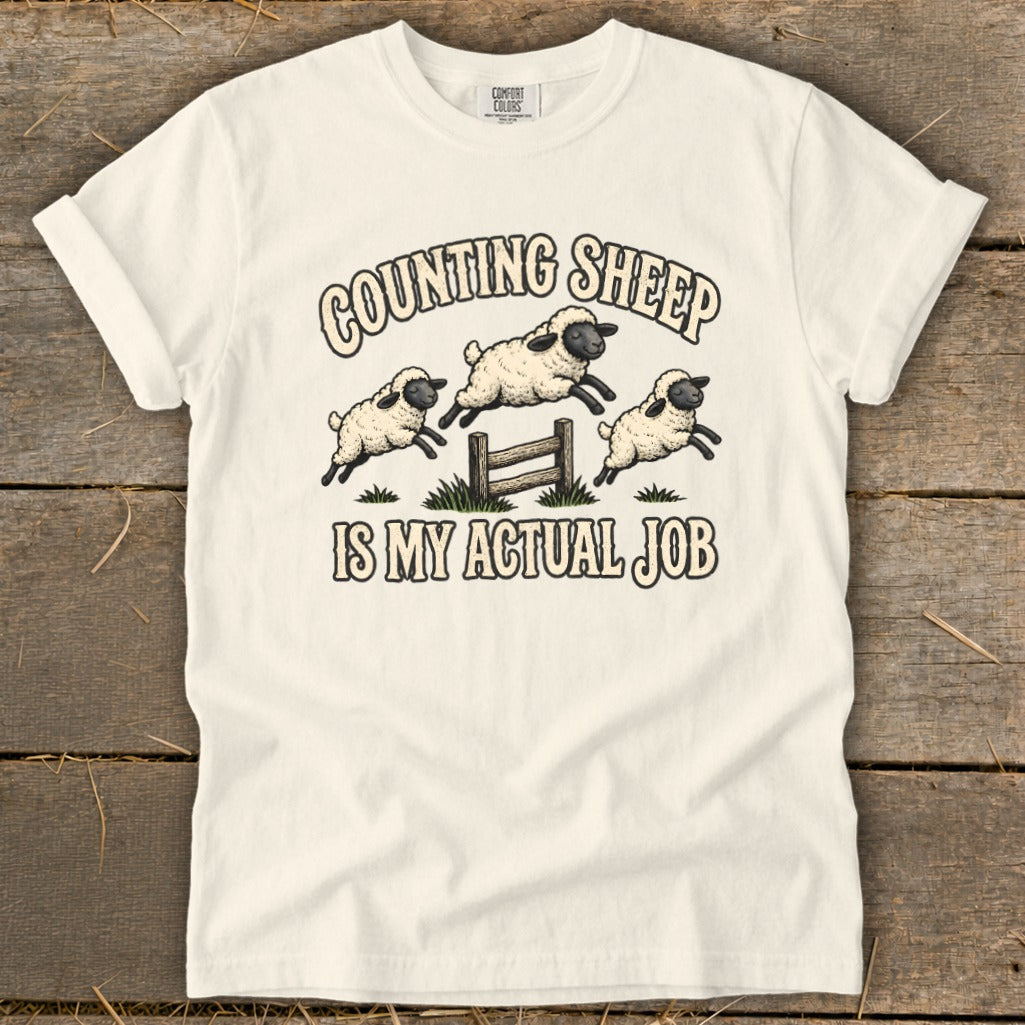 White t-shirt with "Counting Sheep Is My Actual Job" text and jumping sheep graphic.