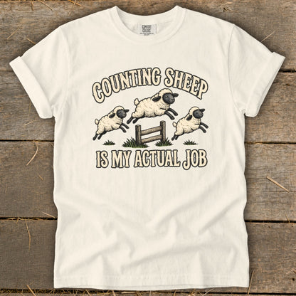White t-shirt with "Counting Sheep Is My Actual Job" text and jumping sheep graphic.