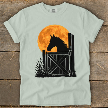 Light green t-shirt featuring a graphic of a horse looking out a stable gate against a large orange moon.