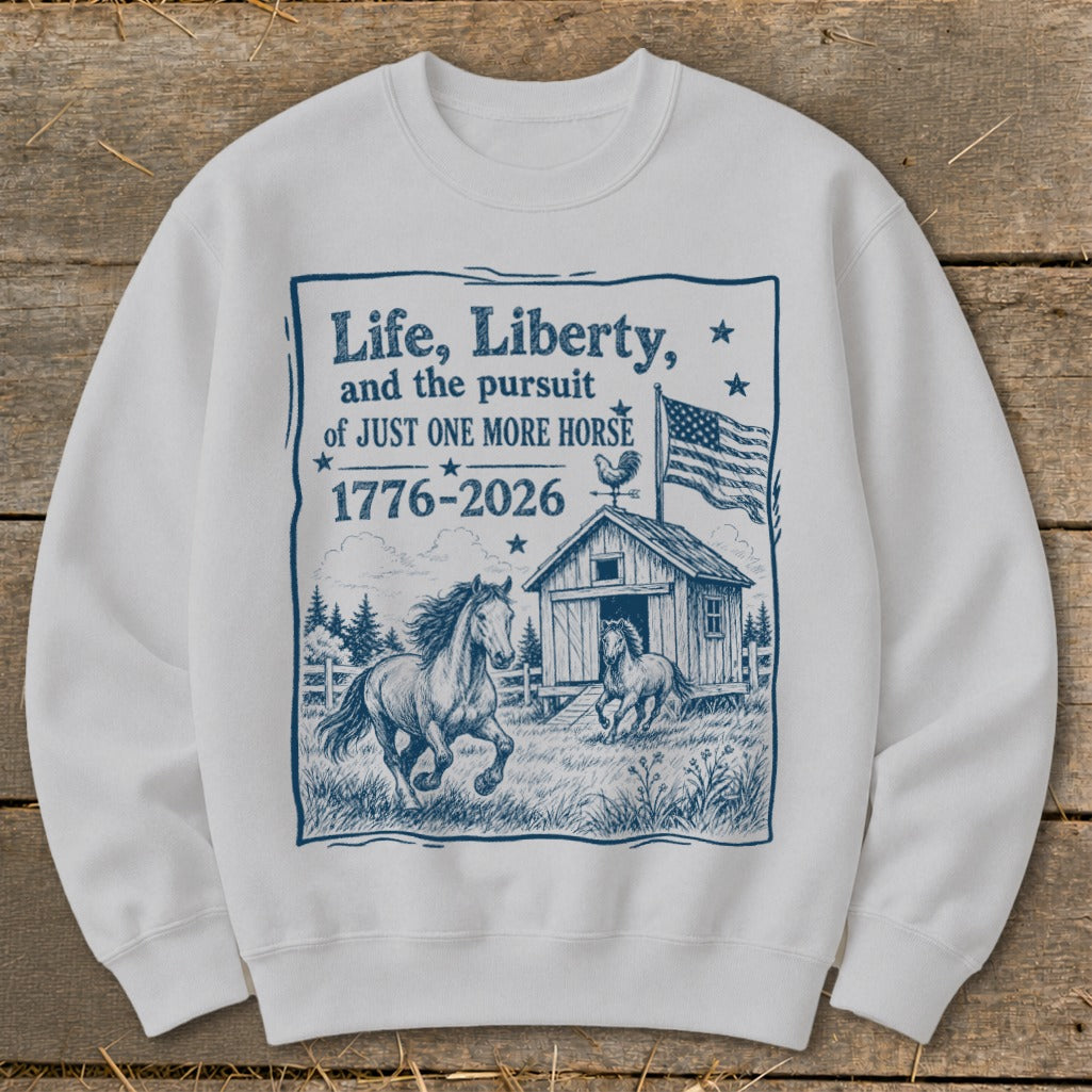 White sweatshirt with blue graphic of horses, barn, flag, and text "Life, Liberty, and the pursuit of JUST ONE MORE HORSE 1776-2026".