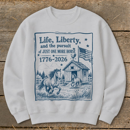 White sweatshirt with blue graphic of horses, barn, flag, and text "Life, Liberty, and the pursuit of JUST ONE MORE HORSE 1776-2026".
