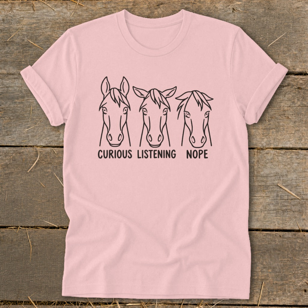 Light pink t-shirt featuring line art of three horse heads labeled Curious, Listening, and Nope, lying flat on a wooden surface.