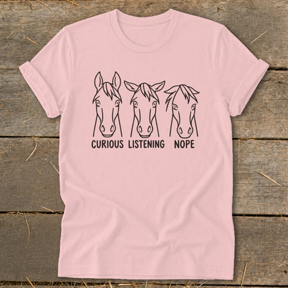 Light pink t-shirt featuring line art of three horse heads labeled Curious, Listening, and Nope, lying flat on a wooden surface.