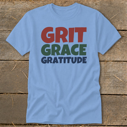 Light blue short-sleeve t-shirt with red, green, and blue text reading GRIT GRACE GRATITUDE, laid flat on a rustic wooden surface.