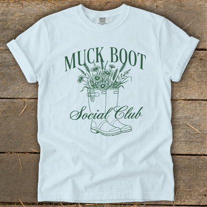 White t-shirt with green text and graphic of muck boots filled with flowers.