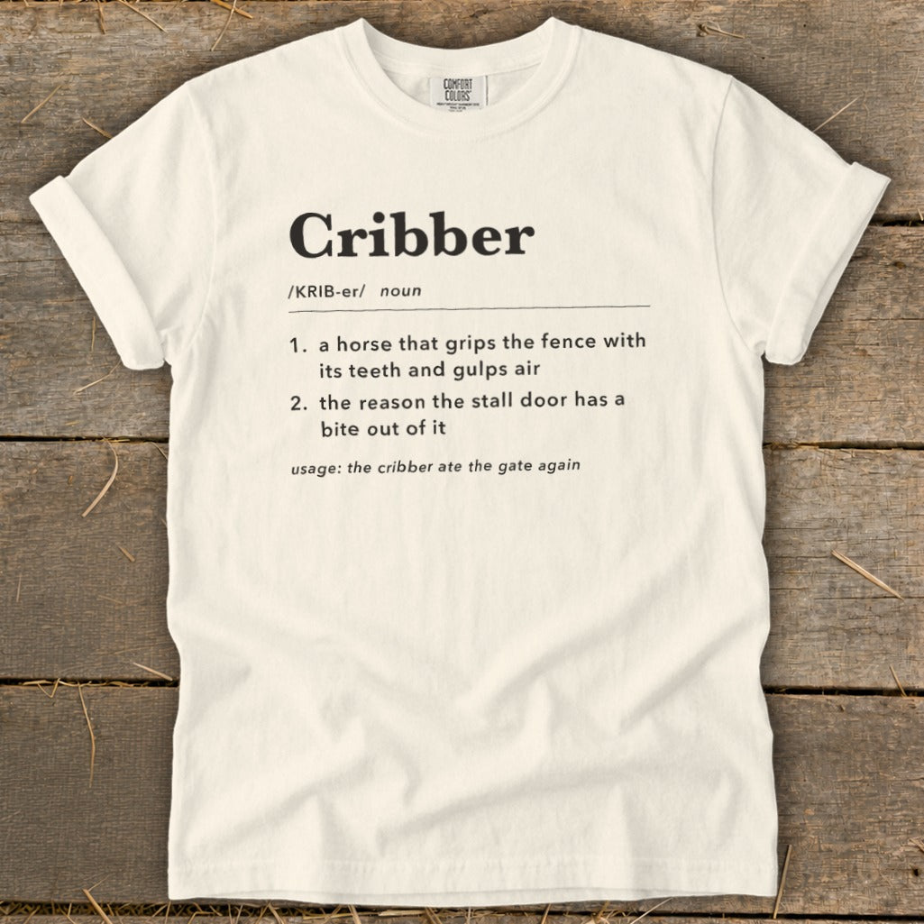 A cream-colored t-shirt with black dictionary definition text for the word Cribber printed on the chest.
