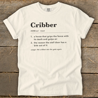 A cream-colored t-shirt with black dictionary definition text for the word Cribber printed on the chest.