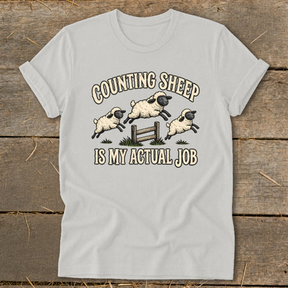 Light grey t-shirt with cartoon sheep jumping over a fence with text "COUNTING SHEEP IS MY ACTUAL JOB".