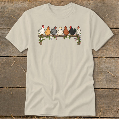 A cream-colored short-sleeve t-shirt featuring a cartoon graphic of seven colorful chickens sitting on a flower-covered wooden fence.