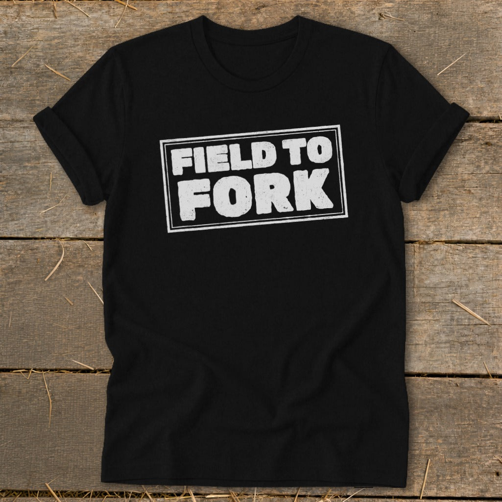 A black crewneck t-shirt with rolled sleeves lies flat on a rustic wooden surface, featuring the white text "FIELD TO FORK" enclosed in a distressed rectangular outline on the chest.