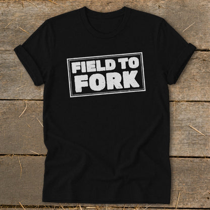 A black crewneck t-shirt with rolled sleeves lies flat on a rustic wooden surface, featuring the white text "FIELD TO FORK" enclosed in a distressed rectangular outline on the chest.
