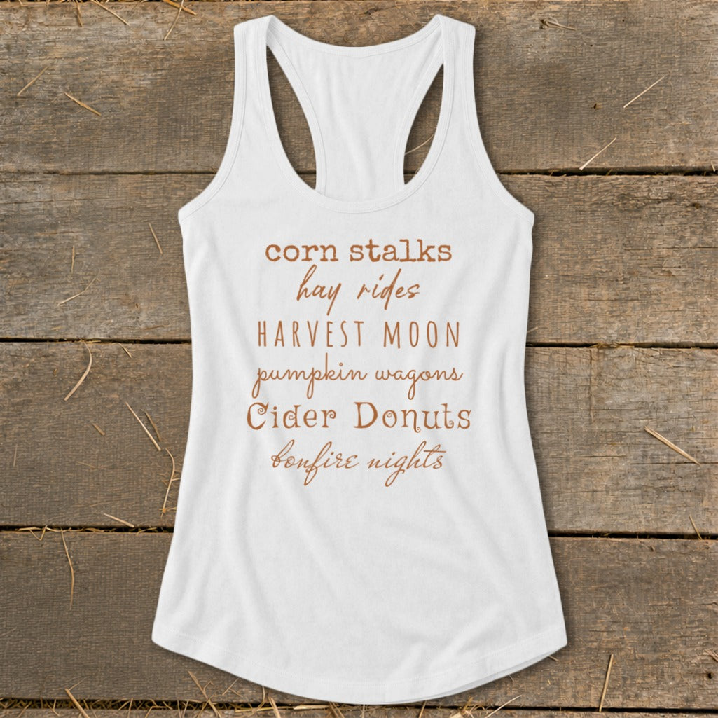 White tank top with text listing fall activities: corn stalks, hay rides, harvest moon, pumpkin wagons, cider donuts, bonfire nights.