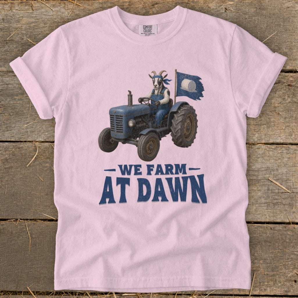 Pink t-shirt graphic of a goat driving a blue tractor, flying a flag. Text reads: WE FARM AT DAWN.