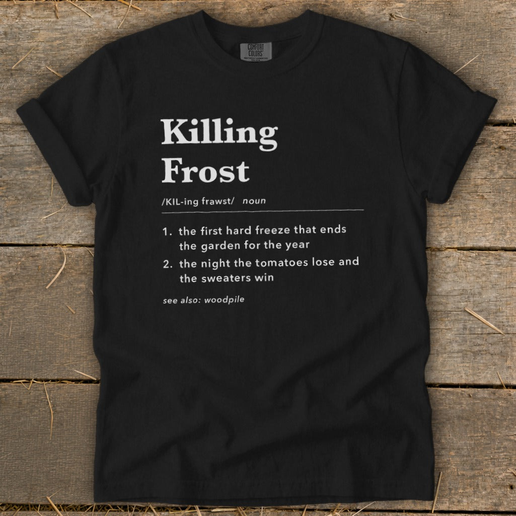 A black t-shirt featuring dictionary-style text defining "Killing Frost" lies flat on a rustic wooden surface.