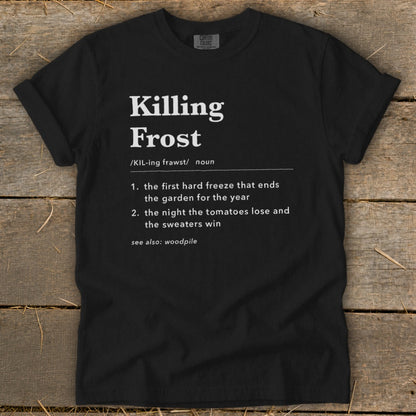 A black t-shirt featuring dictionary-style text defining "Killing Frost" lies flat on a rustic wooden surface.