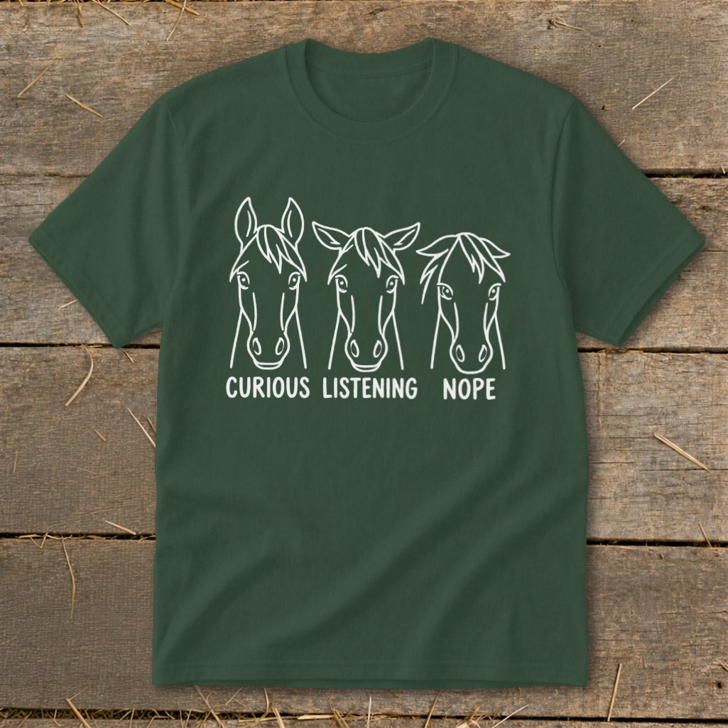 A dark green t-shirt with three white horse line drawings labeled Curious, Listening, and Nope, lying flat on a wooden surface.