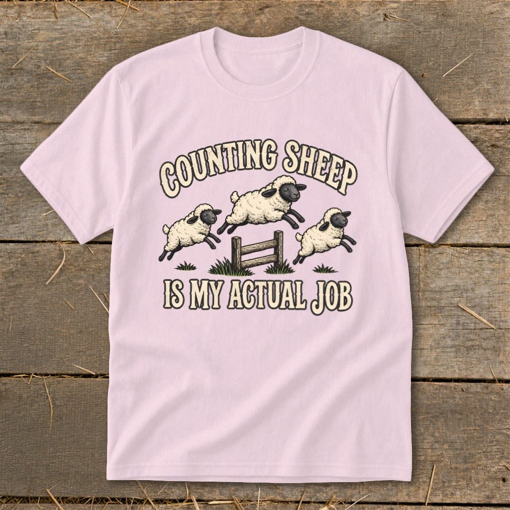 Pink t-shirt with illustration of sheep jumping over a fence and text "COUNTING SHEEP IS MY ACTUAL JOB".