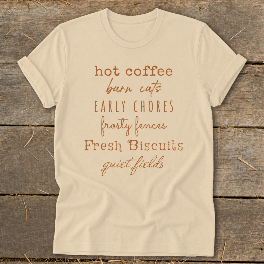 Light tan t-shirt with brown text listing "hot coffee, barn cats, early chores, frosty fences, Fresh Biscuits, quiet fields".