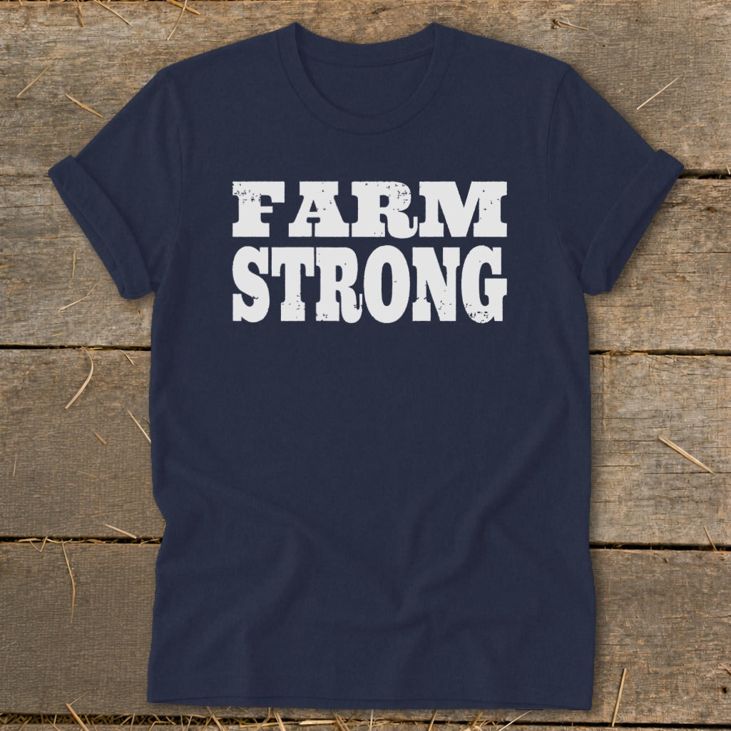 A dark blue crewneck t-shirt with rolled sleeves lies flat on a wooden surface, featuring the white text "FARM STRONG" printed centrally in a distressed, blocky font.
