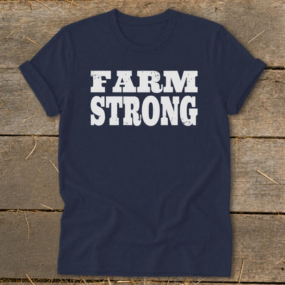 A dark blue crewneck t-shirt with rolled sleeves lies flat on a wooden surface, featuring the white text "FARM STRONG" printed centrally in a distressed, blocky font.