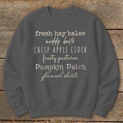 Dark gray sweatshirt with cream text listing autumn activities: fresh hay bales, muddy boots, crisp apple cider, frosty pastures, pumpkin patch, flannel shirts.