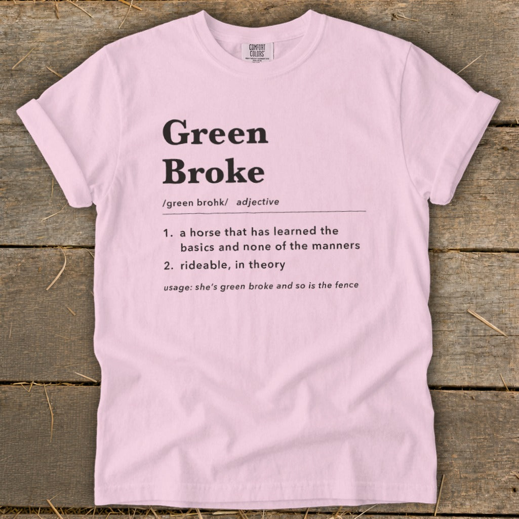 A pink t-shirt printed with a dictionary-style definition of Green Broke on a rustic wooden background.