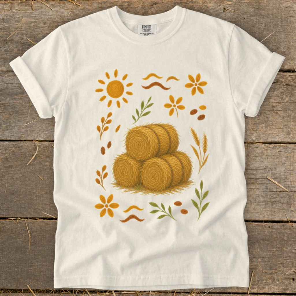 White t-shirt with fall harvest design featuring hay bales, sun, and leaves.