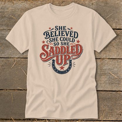 Light tan t-shirt with "She believed she could so she saddled up" text and horseshoe.