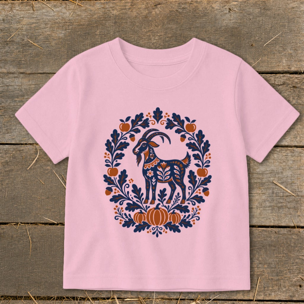 A light pink t-shirt displays a folk art goat surrounded by autumn leaves, pumpkins, and acorns.