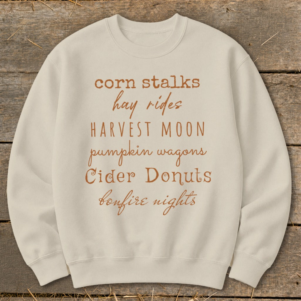Light beige crewneck sweatshirt with fall-themed text list: corn stalks, hay rides, harvest moon, pumpkin wagons, cider donuts, bonfire nights.