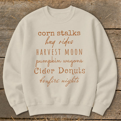 Light beige crewneck sweatshirt with fall-themed text list: corn stalks, hay rides, harvest moon, pumpkin wagons, cider donuts, bonfire nights.