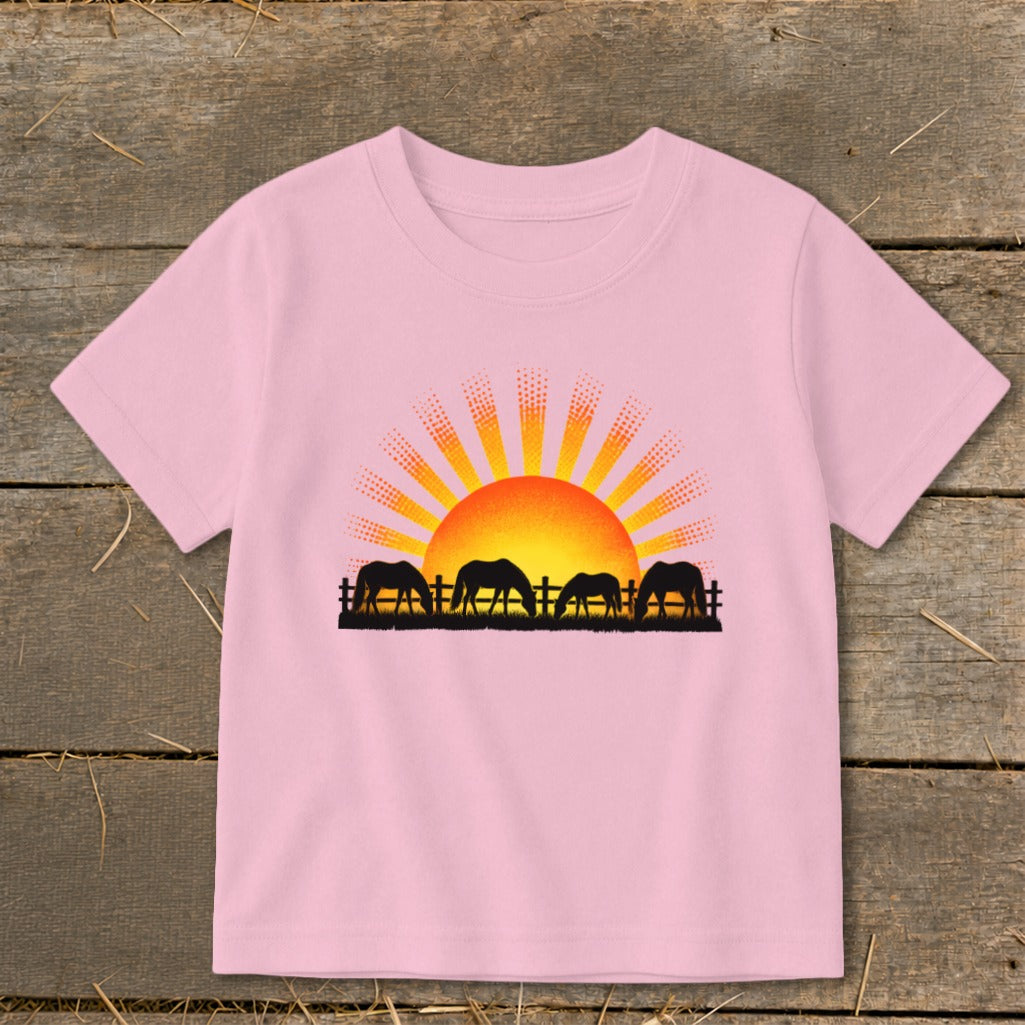 Pink t-shirt with a sunset graphic and silhouette of horses grazing behind a fence.