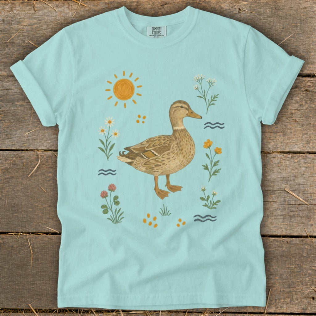 Duck surrounded by flowers, sun, and water waves on a light blue t-shirt.