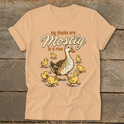 Peach t-shirt with illustration of a mother duck and ducklings, text reads "my ducks are Mostly in a row".