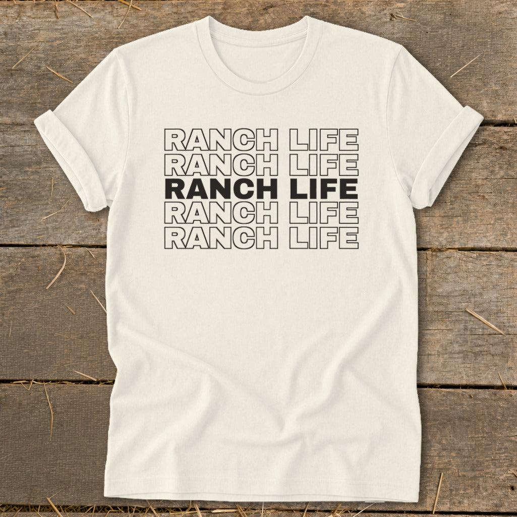 A cream-colored crewneck t-shirt with rolled sleeves laid flat on a rustic wooden surface, featuring the repeated black text "RANCH LIFE" stacked in five rows across the chest.