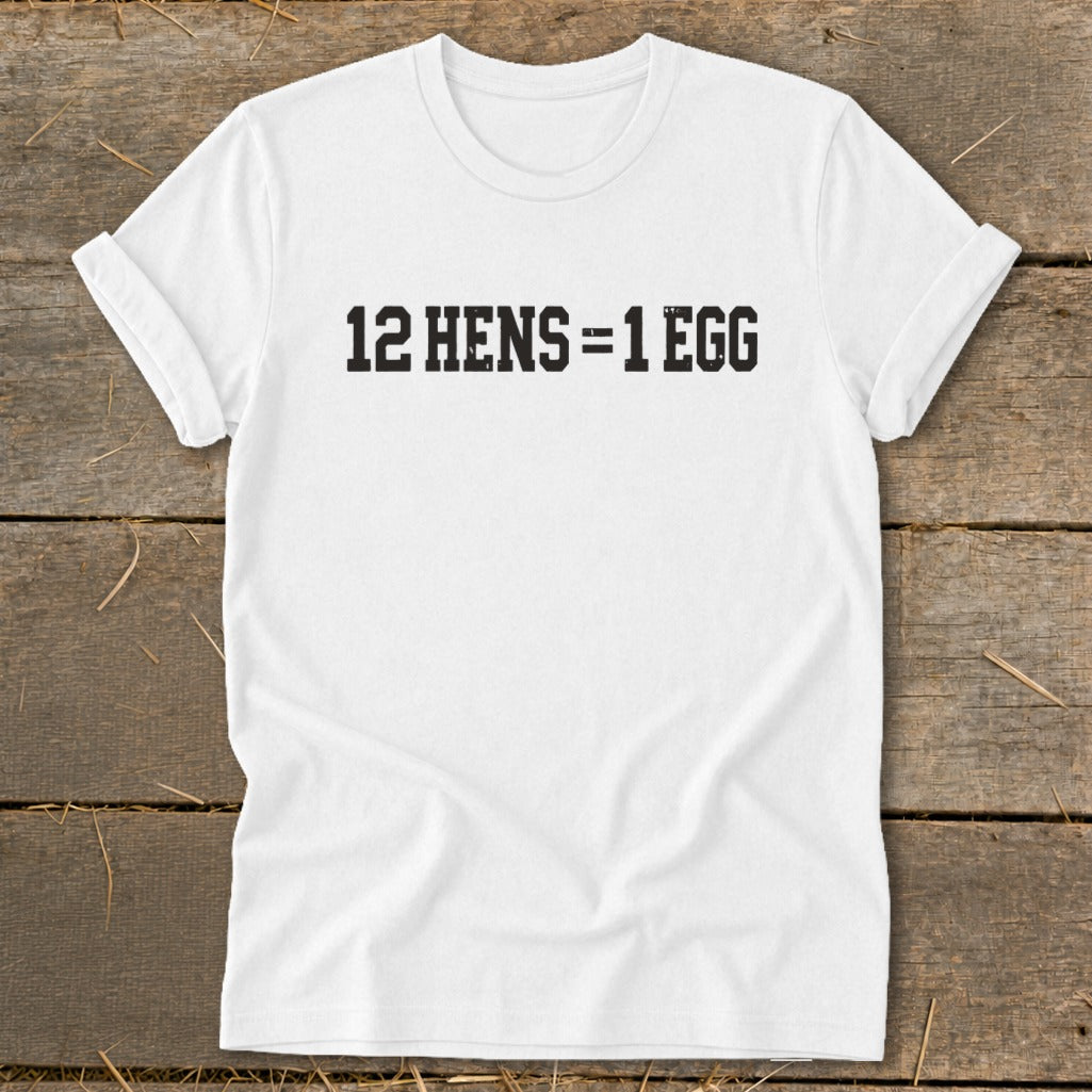 A white t-shirt laid flat on a wooden surface, featuring the black text 12 HENS = 1 EGG.