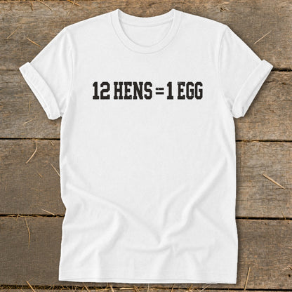 A white t-shirt laid flat on a wooden surface, featuring the black text 12 HENS = 1 EGG.