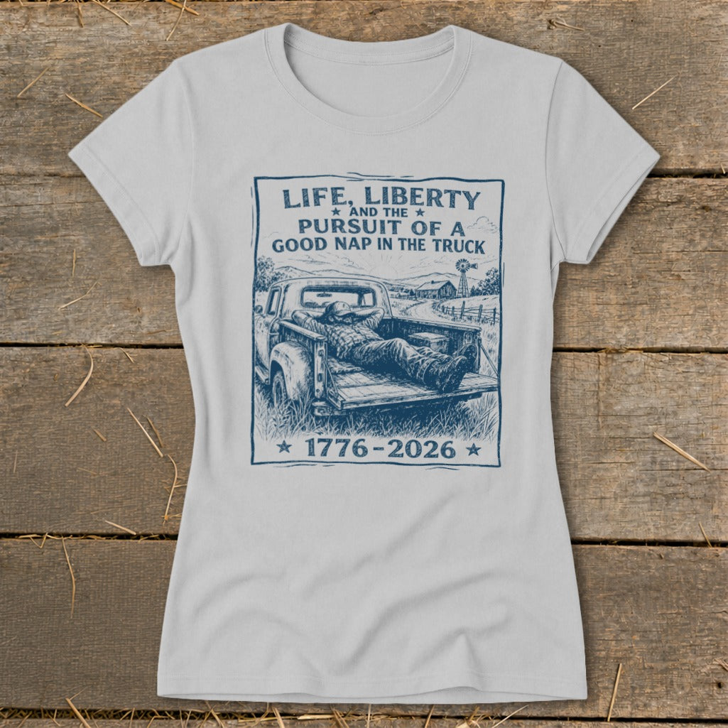 Light grey t-shirt with a sketch of a man napping in a truck bed, text reads "Life, Liberty and the Pursuit of a Good Nap in the Truck 1776-2026".