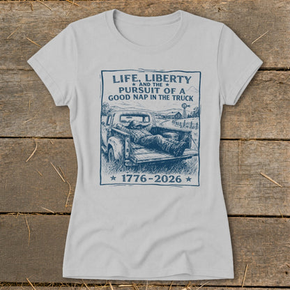 Light grey t-shirt with a sketch of a man napping in a truck bed, text reads "Life, Liberty and the Pursuit of a Good Nap in the Truck 1776-2026".