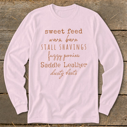 Pink long-sleeve shirt with text: sweet feed, warm barn, STALL SHAVINGS, fuzzy ponies, Saddle Leather, dusty boots.