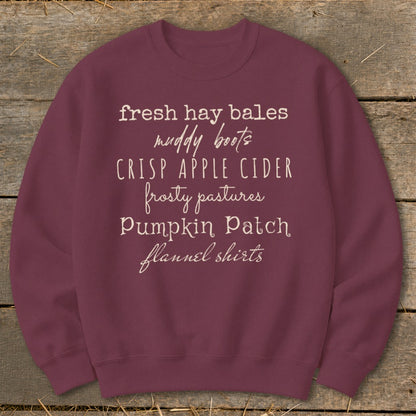 Burgundy sweatshirt with white text listing fall themes: fresh hay bales, muddy boots, crisp apple cider, frosty pastures, pumpkin patch, flannel shirts.