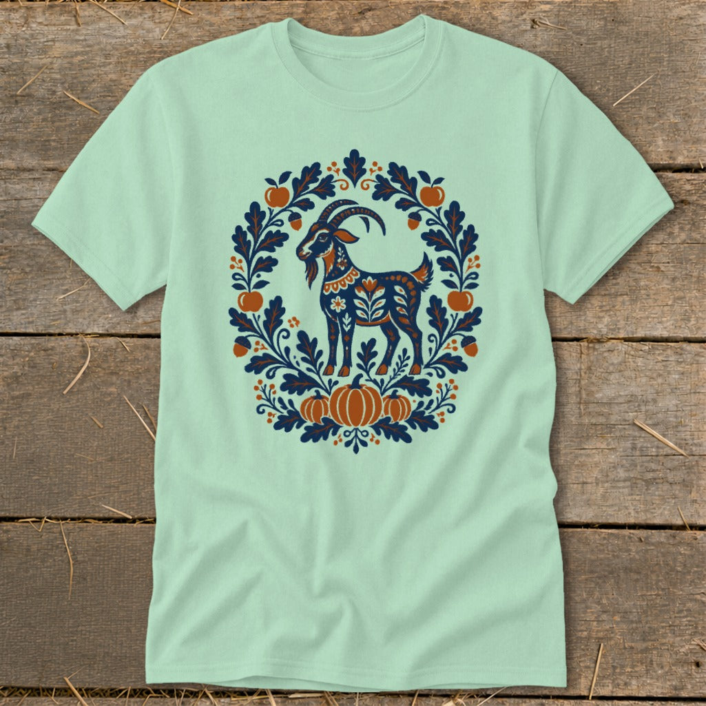 A light green short-sleeved t-shirt features a folkloric design of a goat inside a floral wreath.