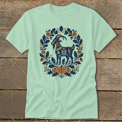 A light green short-sleeved t-shirt features a folkloric design of a goat inside a floral wreath.