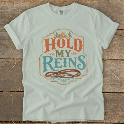 Light blue t-shirt with "Hold My Reins" text, cacti, sun, and leather reins graphic.