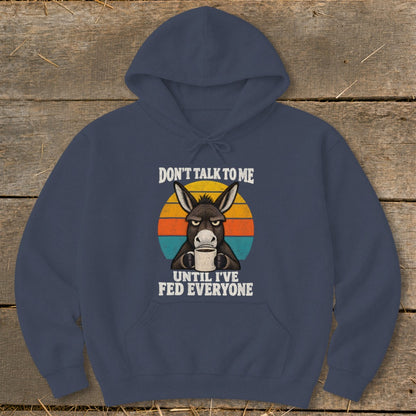 A dark blue hoodie featuring a grumpy donkey holding a coffee mug in front of a retro sunset graphic with the text DON'T TALK TO ME UNTIL I'VE FED EVERYONE.