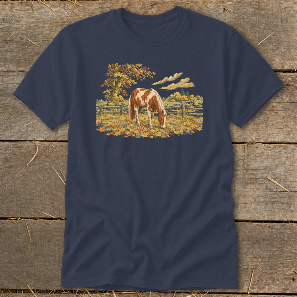 Brown and white horse grazing in a field of autumn leaves under a tree.