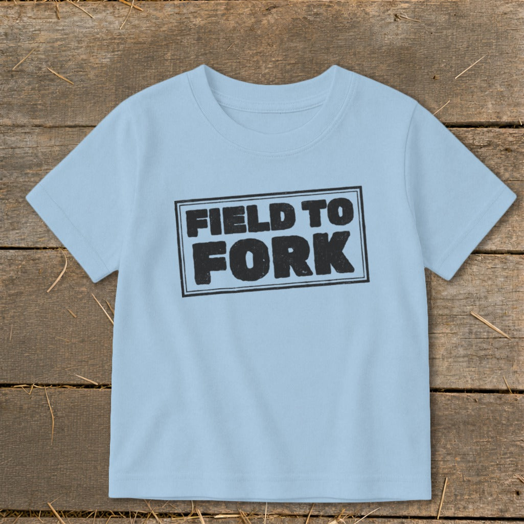 Light blue children's t-shirt with a black boxed text graphic reading Field to Fork, laid flat on a rustic wooden surface.