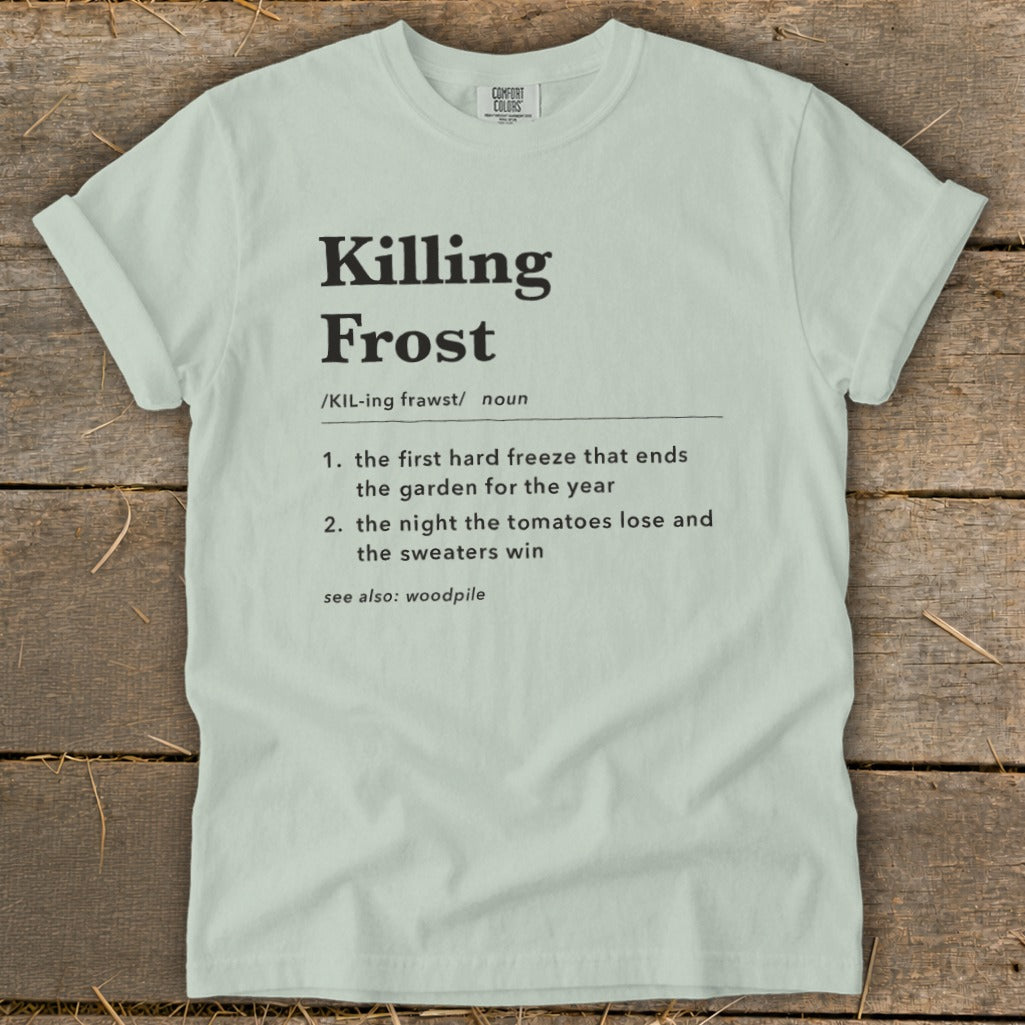 A light green t-shirt with black dictionary-style text defining the phrase Killing Frost laid flat on wooden planks.