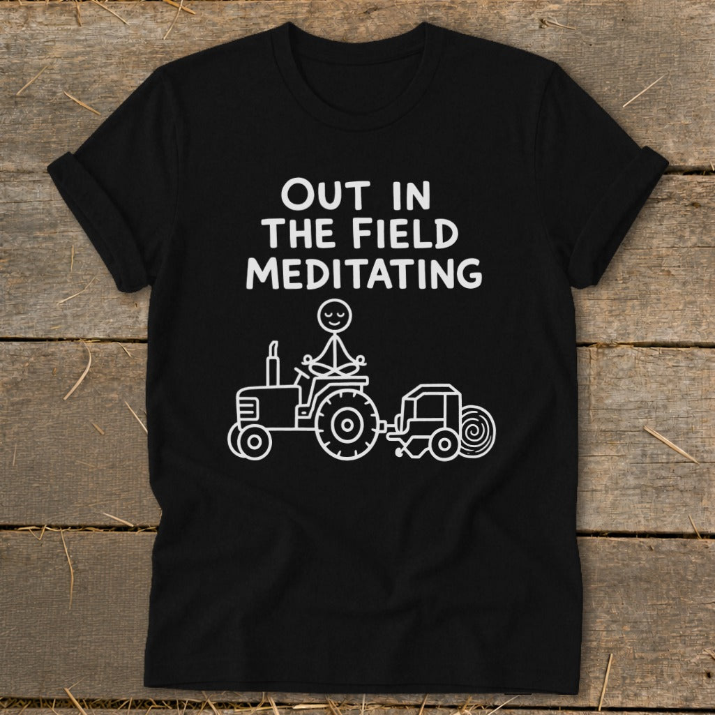 Black t-shirt with white text "Out in the field meditating" and stick figure on a tractor.